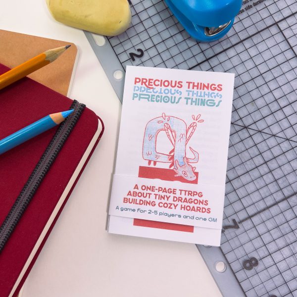 A small 1/8th of a letter sheet of paper sized book titled Precious Things sits on top of a gridded cutting mat surrounded by stationery, pencils, notebooks, a small eraser and a small stapler.